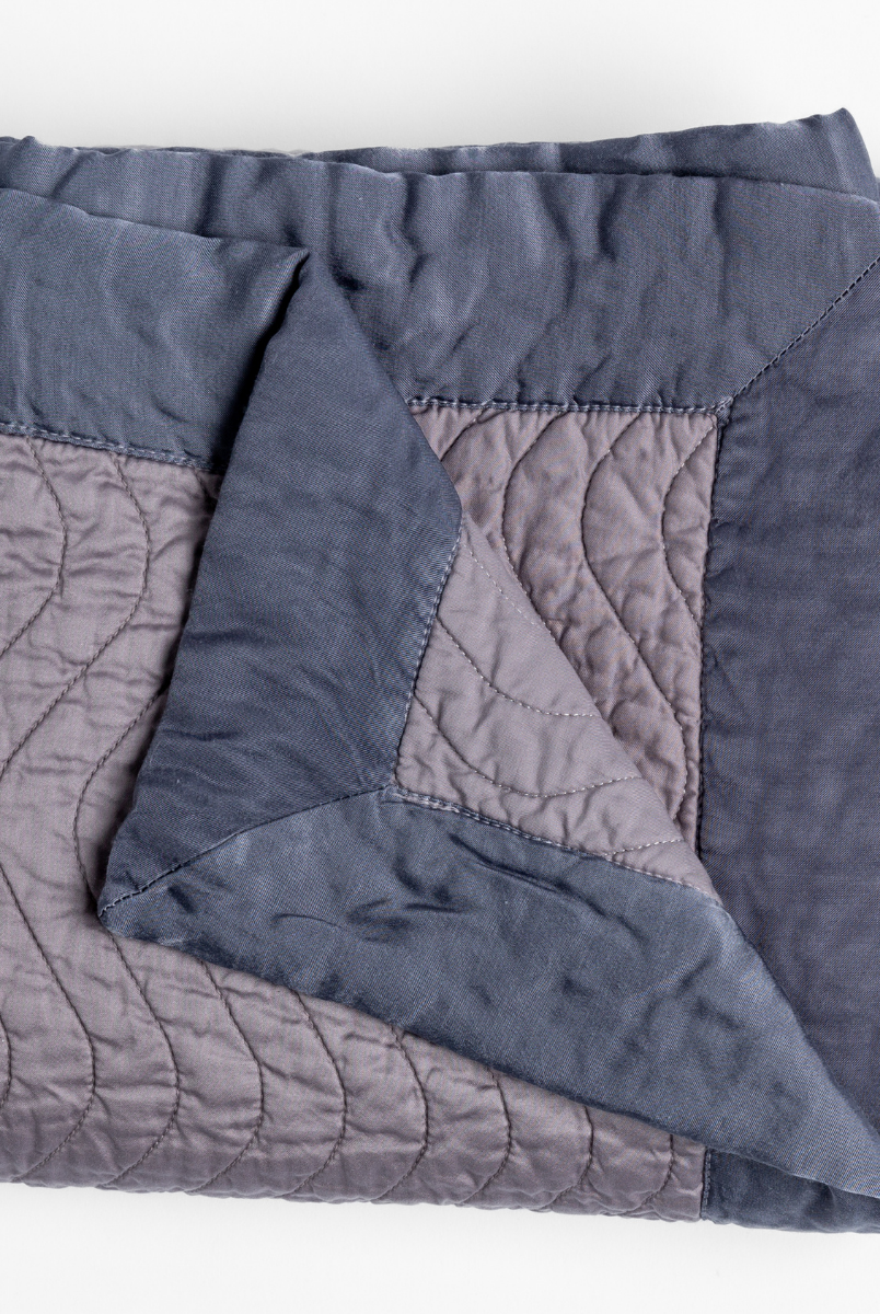 French Lavender: a folded quilted cotton sateen throw blanket with its corner folded down to show the trim contrast - shot against a white background. 