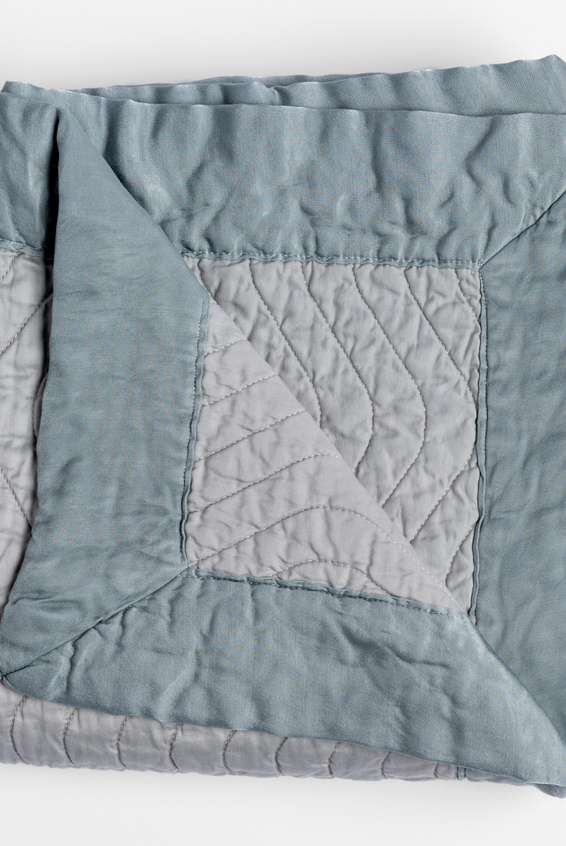 Mineral: a folded quilted cotton sateen throw blanket with its corner folded down to show the trim contrast - shot against a white background. 