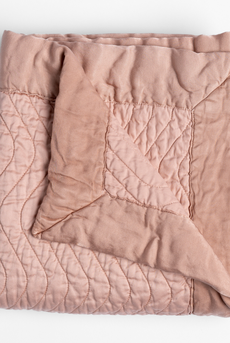 Rouge: a folded quilted cotton sateen throw blanket with its corner folded down to show the trim contrast - shot against a white background. 